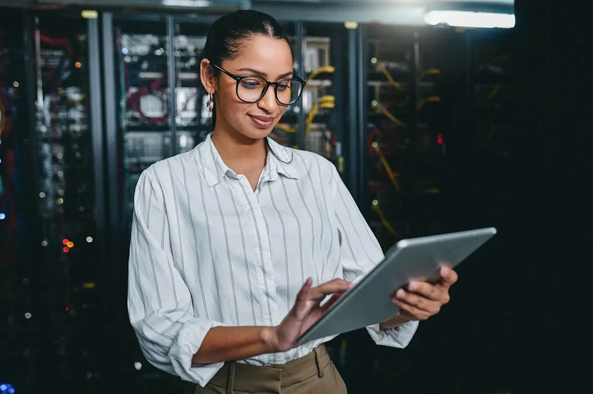 A woman in a striped shirt using a tablet in a data center filled with server racks.