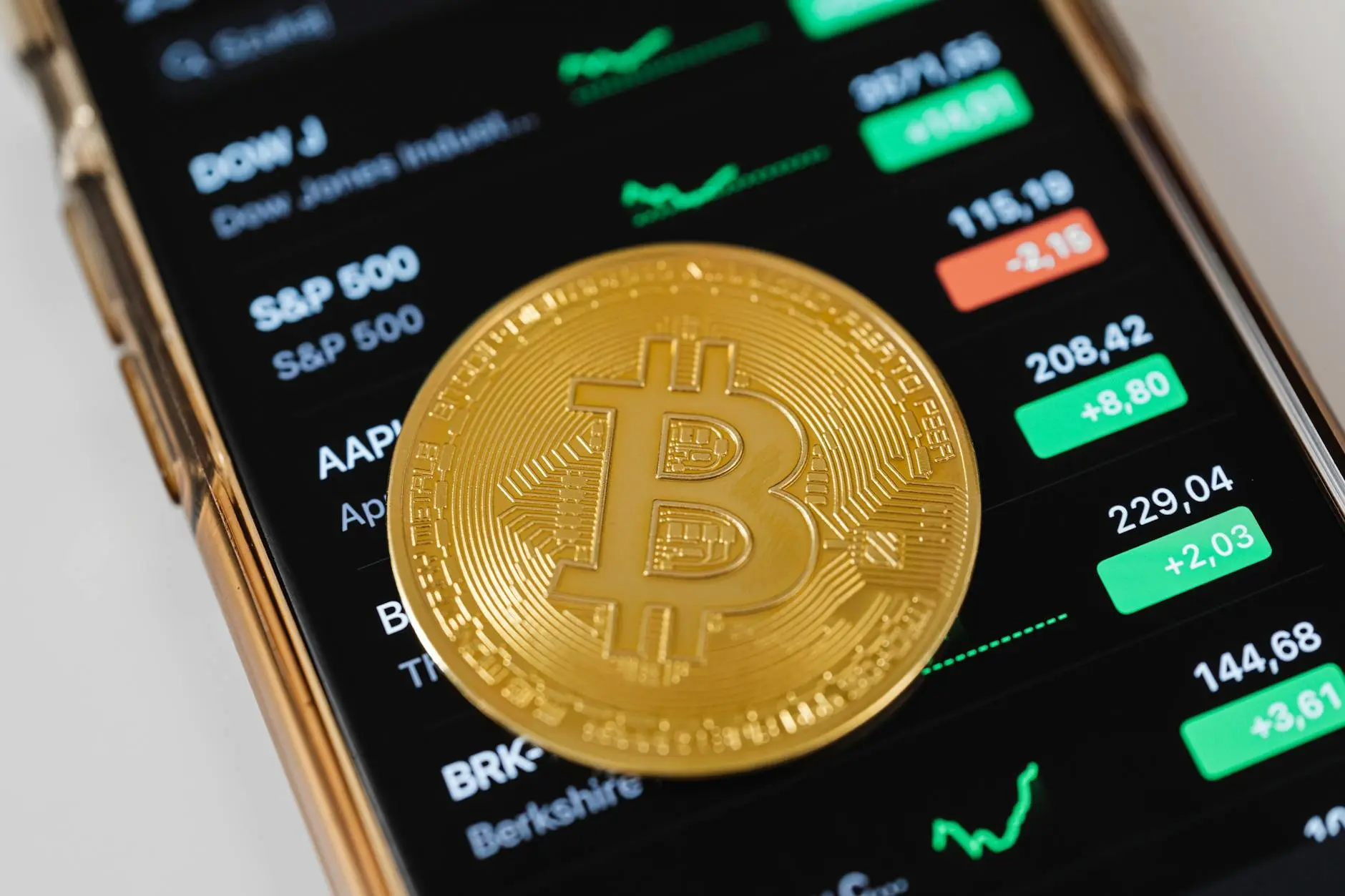 A Bitcoin coin on top of a smartphone screen showing stock market data.