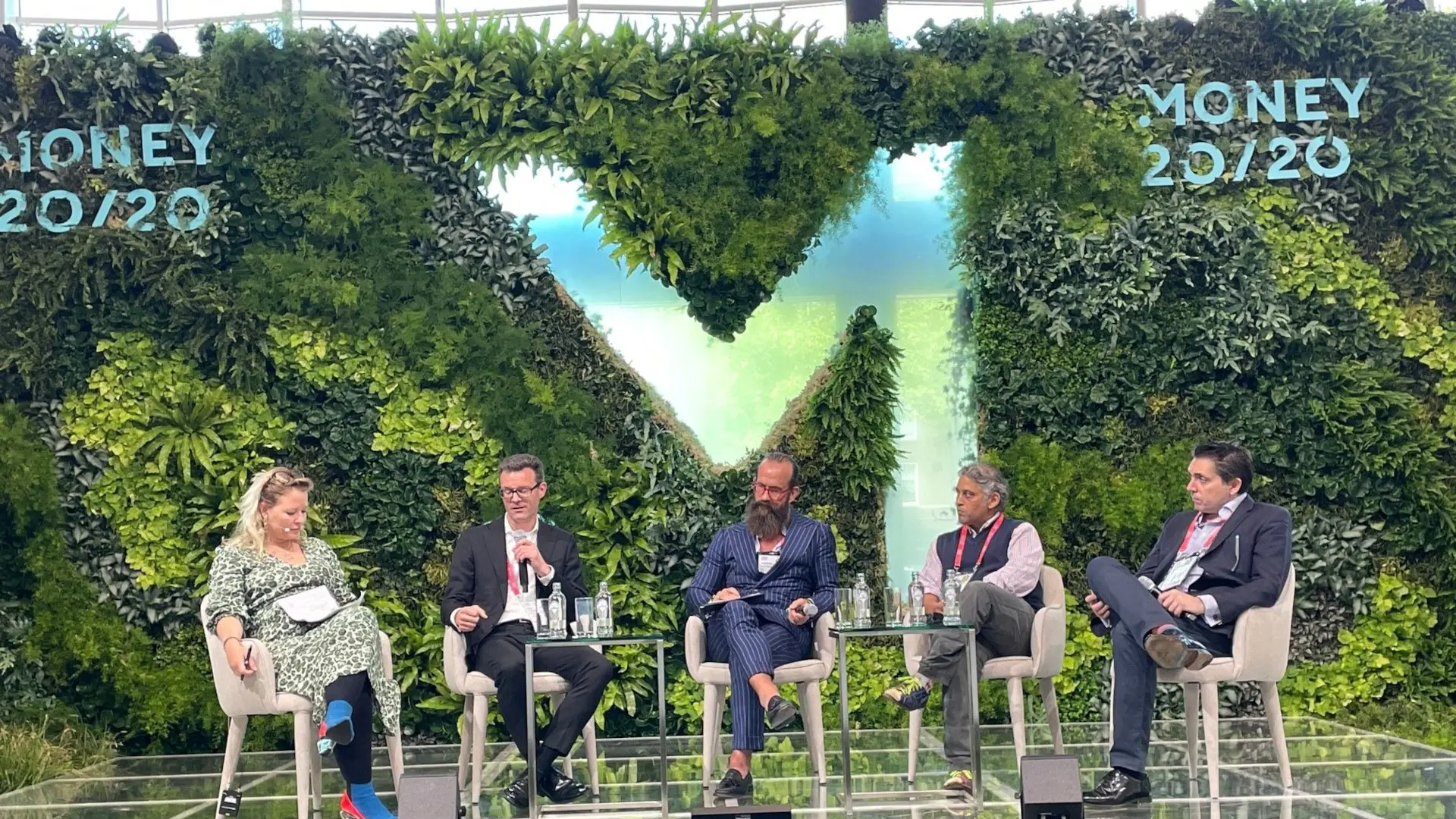 Eine Diskussionsrunde auf der Bühne der Money20/20 Konferenz vor einer grünen Pflanzenwand.